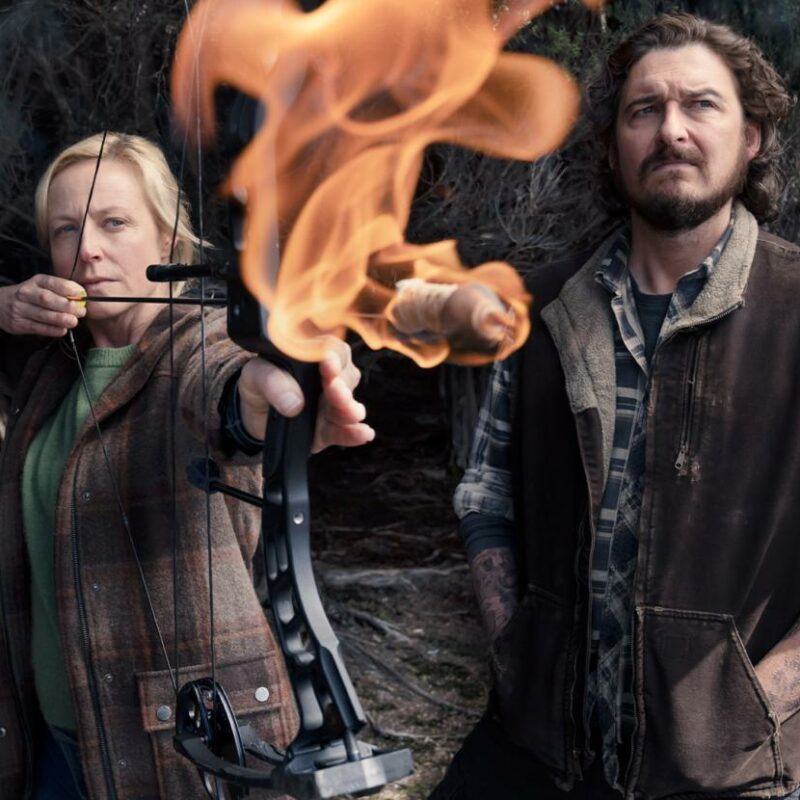 An assured blonde woman (actor Marta Dusseldorp as Stella Heikkinen) stands with a flaming crossbow pulled tautly, beside her a laconic, rural man (acor Toby Leonard Moore, as Jeremiah) wearing a plaid long sleeve shirt and brown coat for warmth, looks out to the distance.